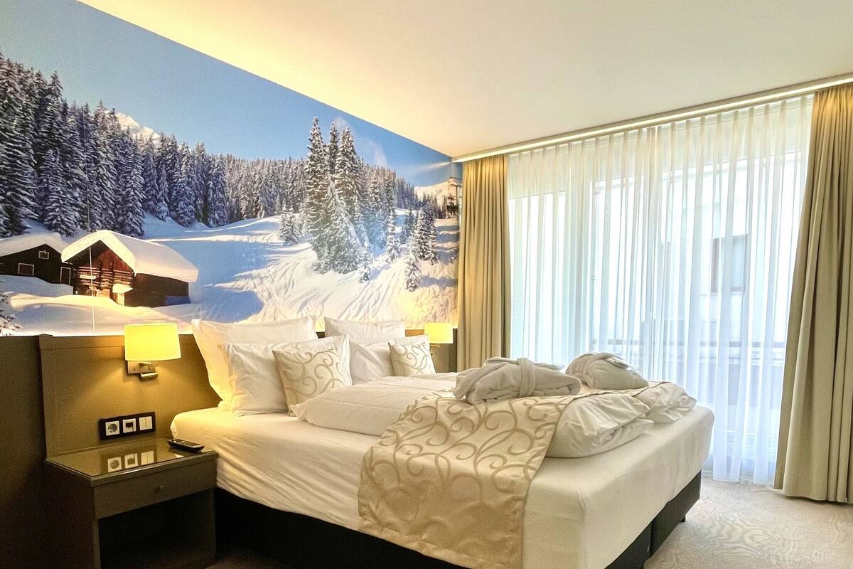 Hotelkamer met een groot bed, nachtkastje, lamp en muurschildering van een besneeuwd berglandschap.