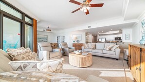 A spacious living room with a large window, a ceiling fan, and a view of the kitchen. - New to the Rental Market, New Owner, Beachfront!! (Orange Beach)