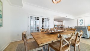 A spacious dining area with a wooden table and chairs, a kitchen in the background, and a living area with a sofa and a painting on the wall. - New to the Rental Market, New Owner, Beachfront!! (Orange Beach)