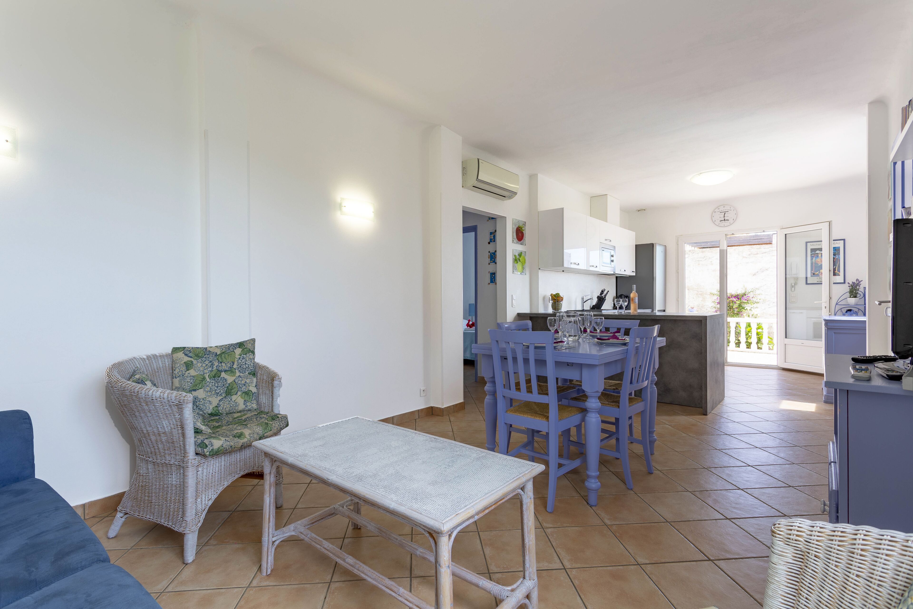 A modern living room with a blue sofa, a wicker chair, and a small table. A kitchen with a dining area is visible in the background.