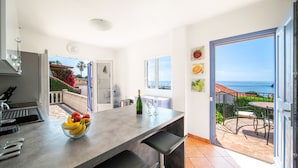 A modern kitchen with a fruit bowl, wine bottle, and glasses on the counter. A sliding door leads to a patio with a table and chairs. - Villa La Cigalette-Gaia's Villas,Beachside Estate with Pool-Steps from the Beach (Théoule-sur-Mer)