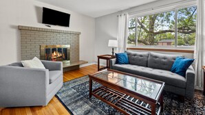 A living room with a fireplace, a grey sofa, a coffee table, and a large window. - Charming Arvada Home - Walking distance to Olde Town (Arvada)