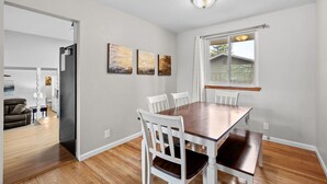 A dining area with a wooden table and white chairs, a window with curtains, and three paintings on the wall. - Charming Arvada Home - Walking distance to Olde Town (Arvada)