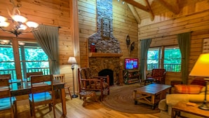 Ein gemütliches Blockhaus mit Holzwänden, einem Steinkamin, einem Fernseher und einem Essbereich mit Holztischen und -stühlen.