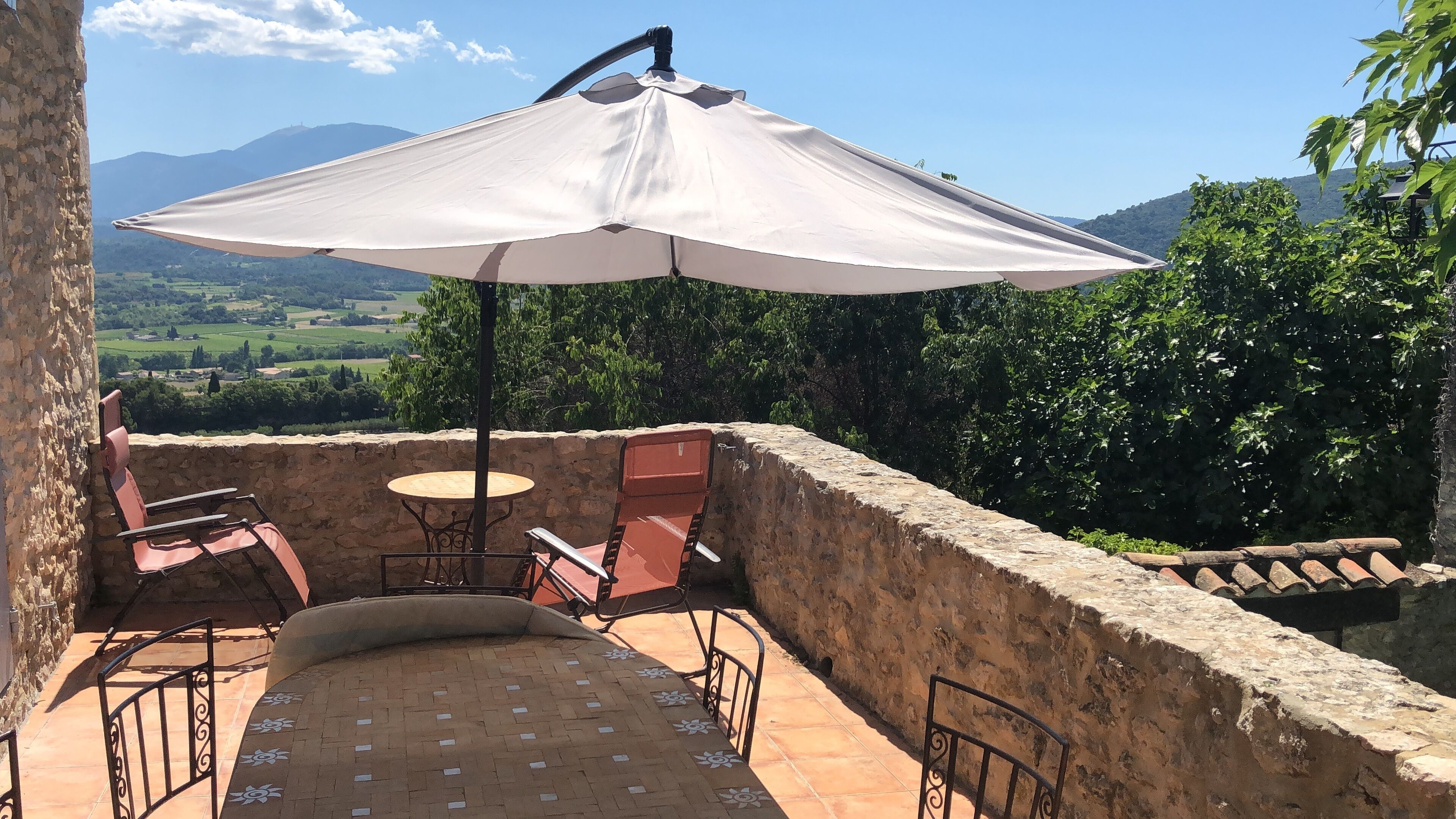 A patio with a table and chairs, a large umbrella, and a view of a green landscape with mountains in the distance.