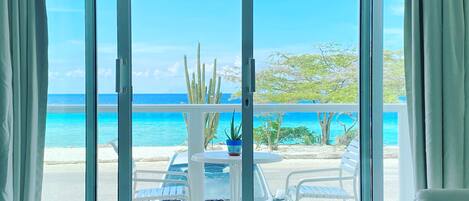 A room with a sliding glass door leading to a balcony with a view of the beach and ocean.
