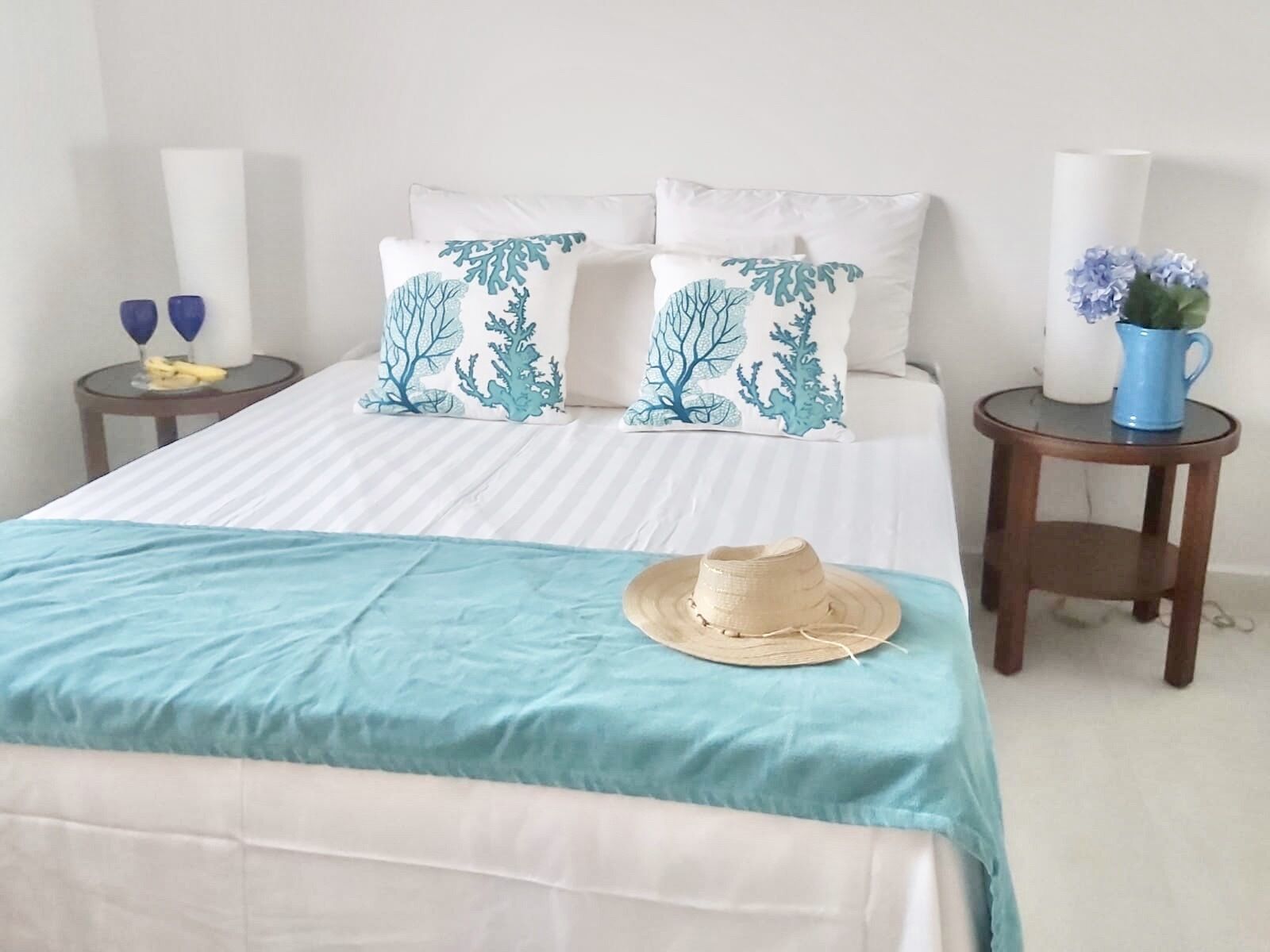 A neatly made bed with a striped bedspread and two decorative pillows, a small round table with a blue mug and a vase of flowers, and a wall-mounted lamp on either side of the bed.