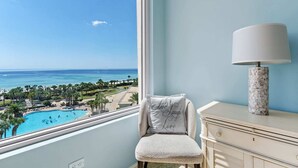 Une chambre avec vue sur une piscine et une plage, une chaise avec un coussin et une lampe sur une commode.