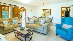 A living room with two sofas, a coffee table, and a blue armchair. There is a dining table with chairs and a chandelier. - GERANIOS. Relax and enjoy! (Puerto Pollensa)