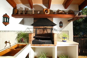A brick-built outdoor kitchen with a grill, a wooden shelf above holding decorative items, and a potted plant on the counter.
