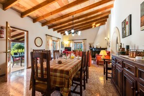 A dining area with a wooden ceiling, a table with a plaid tablecloth, and chairs. There is a fireplace and a sofa in the background.