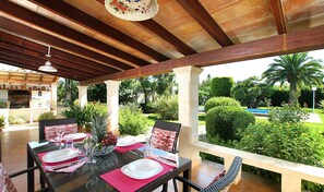 An outdoor patio with a dining table set for two, surrounded by lush greenery and a pool in the background.