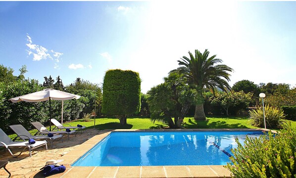 A swimming pool with lounge chairs and umbrellas, surrounded by well-maintained greenery and a clear sky.