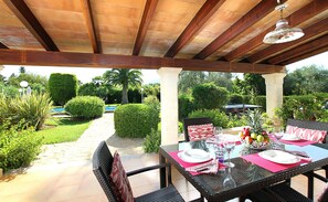 Um pátio externo com mesa posta para refeições, rodeado por vegetação e uma piscina ao fundo.