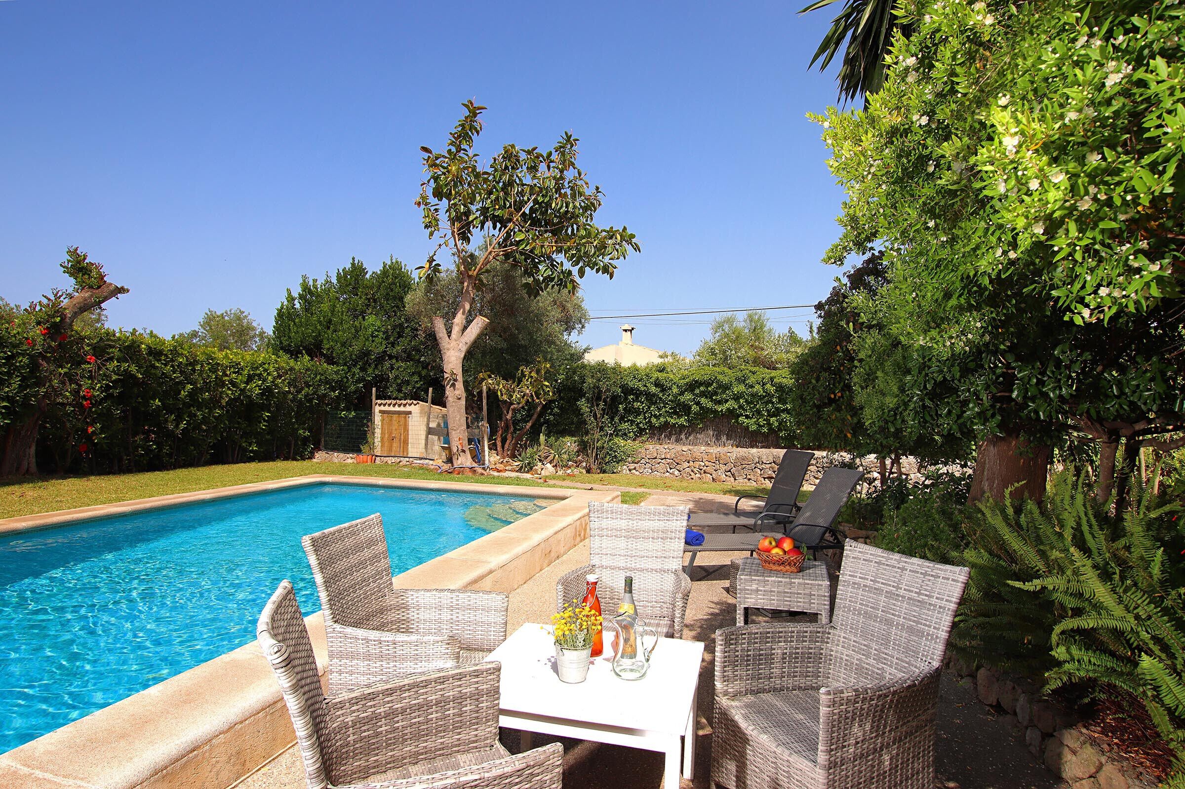 A swimming pool with wicker furniture and a table, surrounded by greenery and a stone wall.