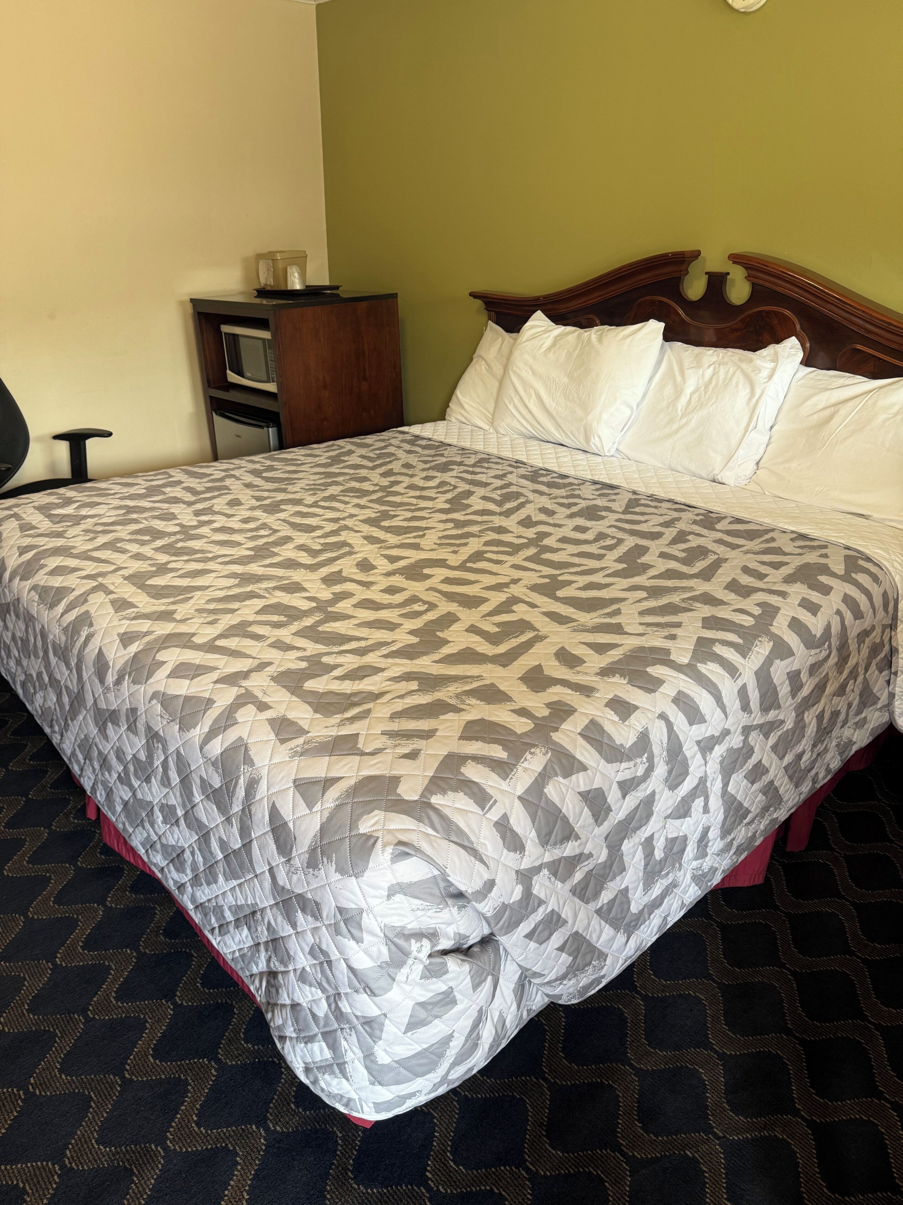 A bed with a patterned bedspread, a wooden headboard, and a small bedside table with a microwave and a refrigerator.