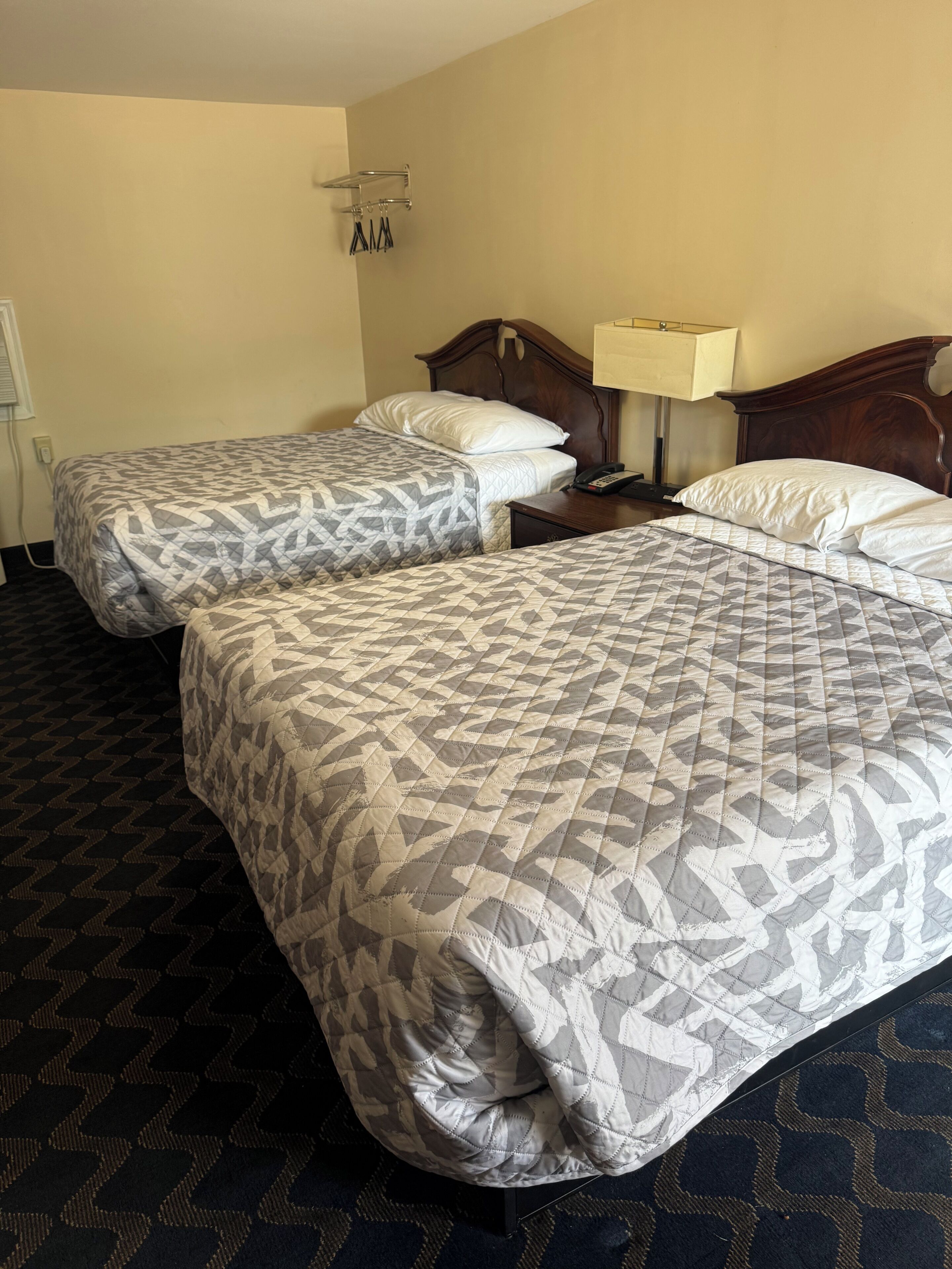 A hotel room with two beds, a nightstand, a lamp, and a wall-mounted towel rack.