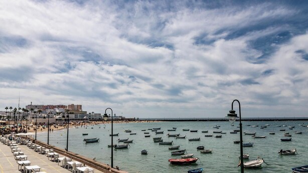 A coastal area with a promenade lined with tables and chairs, boats on the water, and buildings in the background.