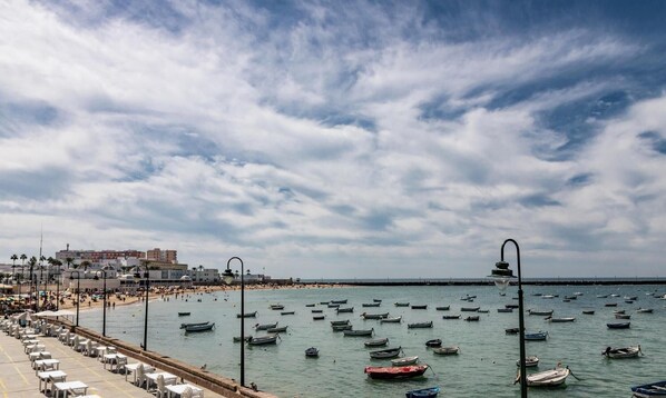 A seaside promenade with rows of white plastic chairs and tables, a beach in the background, and numerous boats on the water.
