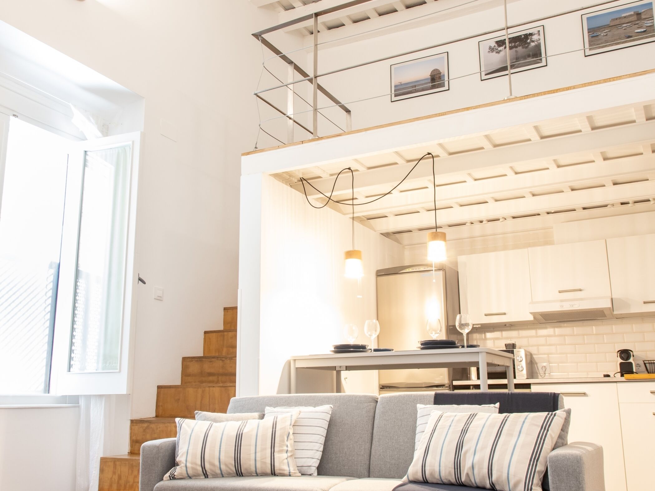 A modern living room with a grey sofa, wooden staircase, and a kitchen area with white cabinets and hanging lights.
