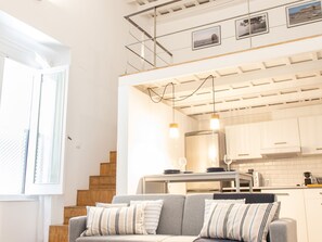 A modern interior with a grey sofa, wooden staircase, white kitchen, and a mezzanine level with framed pictures.