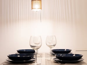 A dining table with four dark bowls and two wine glasses, set against a white background with a hanging light fixture.