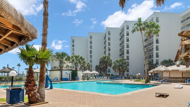 Área de piscina com espreguiçadeiras e guarda-sóis, cercada por palmeiras e um edifício de vários andares.