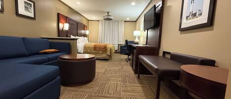 A hotel room with a blue sofa, a round coffee table, a bed with a beige blanket, a desk with a chair, a TV mounted on the wall, and a framed picture of a rocket.