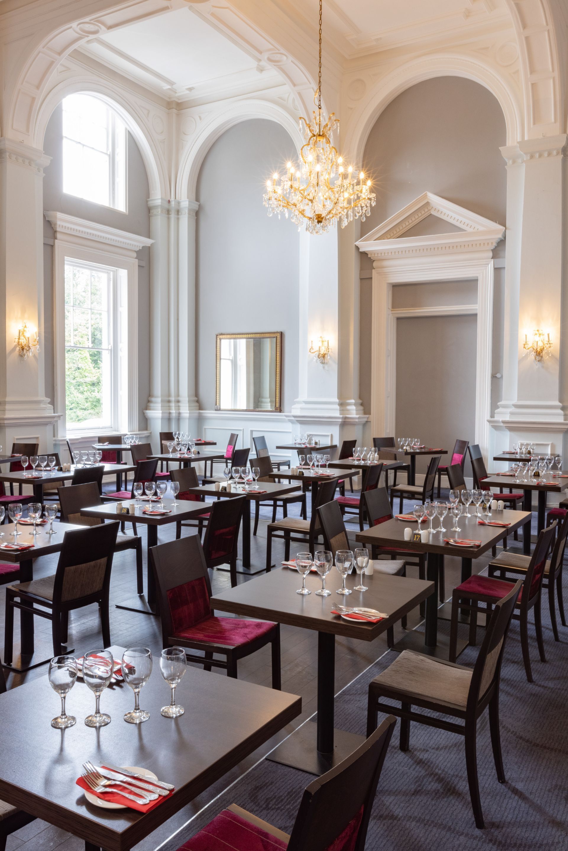 A formal dining room with multiple tables set for service, elegant chandeliers, and classical architecture.