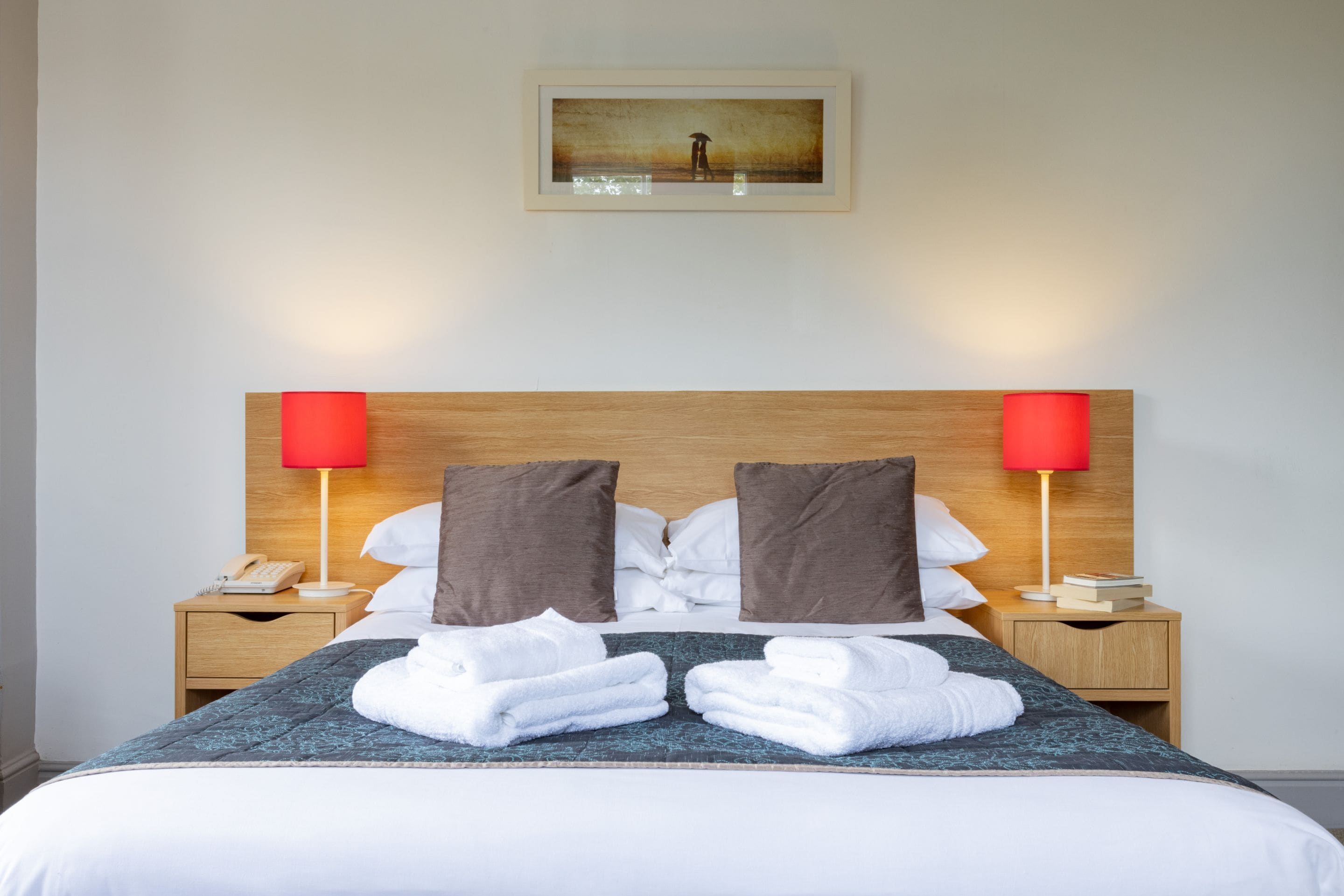 Un lit bien fait, avec deux oreillers, une table de chevet surmontée d’une lampe et un tableau encadré accroché au mur.