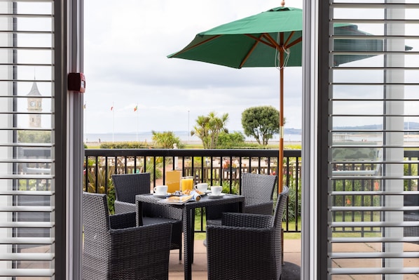 A balcony with wicker chairs and a table set for breakfast, overlooking a scenic view with a clock tower and palm trees. - The Imperial Hotel (Exmouth)