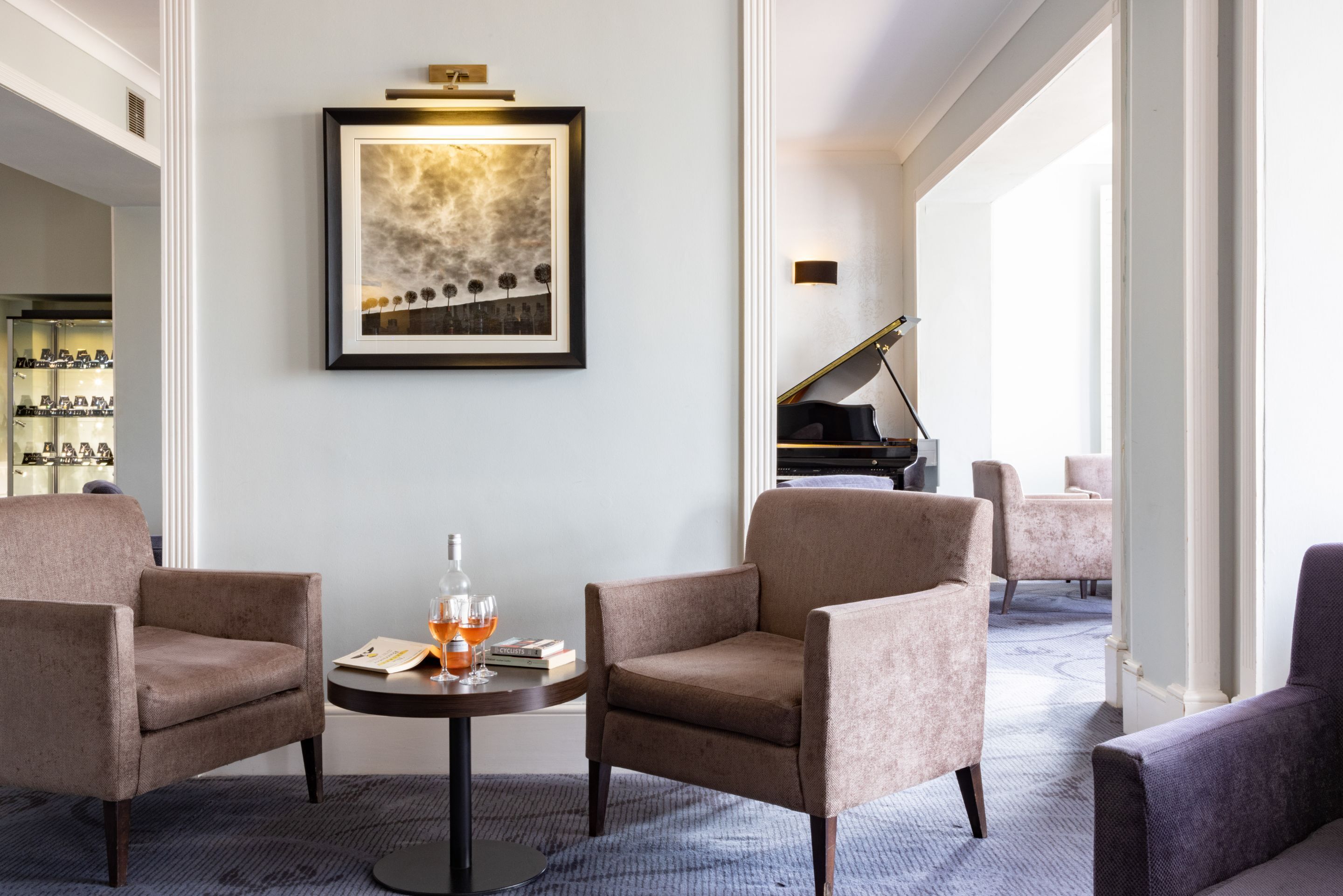 A modern living room with two armchairs, a round table with a drink, a framed abstract painting, and a grand piano in the background.