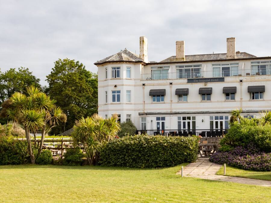 A large, historic building with multiple windows and a central entrance, surrounded by well-maintained gardens and a small pavilion.