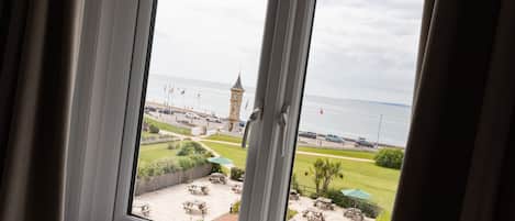 Vista a través de una ventana de una zona costera con una torre del reloj, mesas al aire libre y un césped.