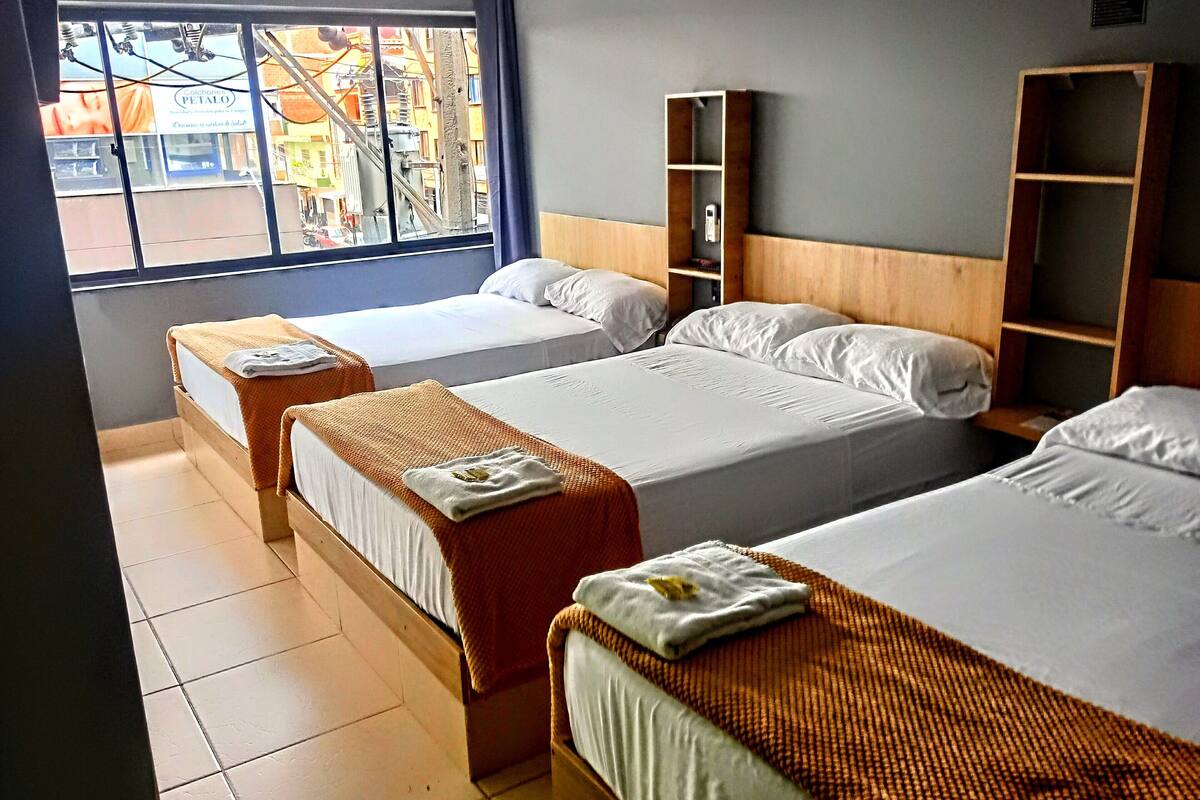 Une chambre d’hôtel avec trois lits simples, une tête de lit en bois, une petite étagère en bois et une fenêtre donnant sur la rue.