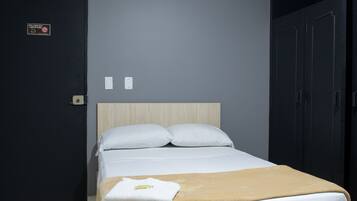 Una camera d'albergo con un letto, una testiera in legno, un cuscino bianco e una coperta beige, con un cartello sulla porta.