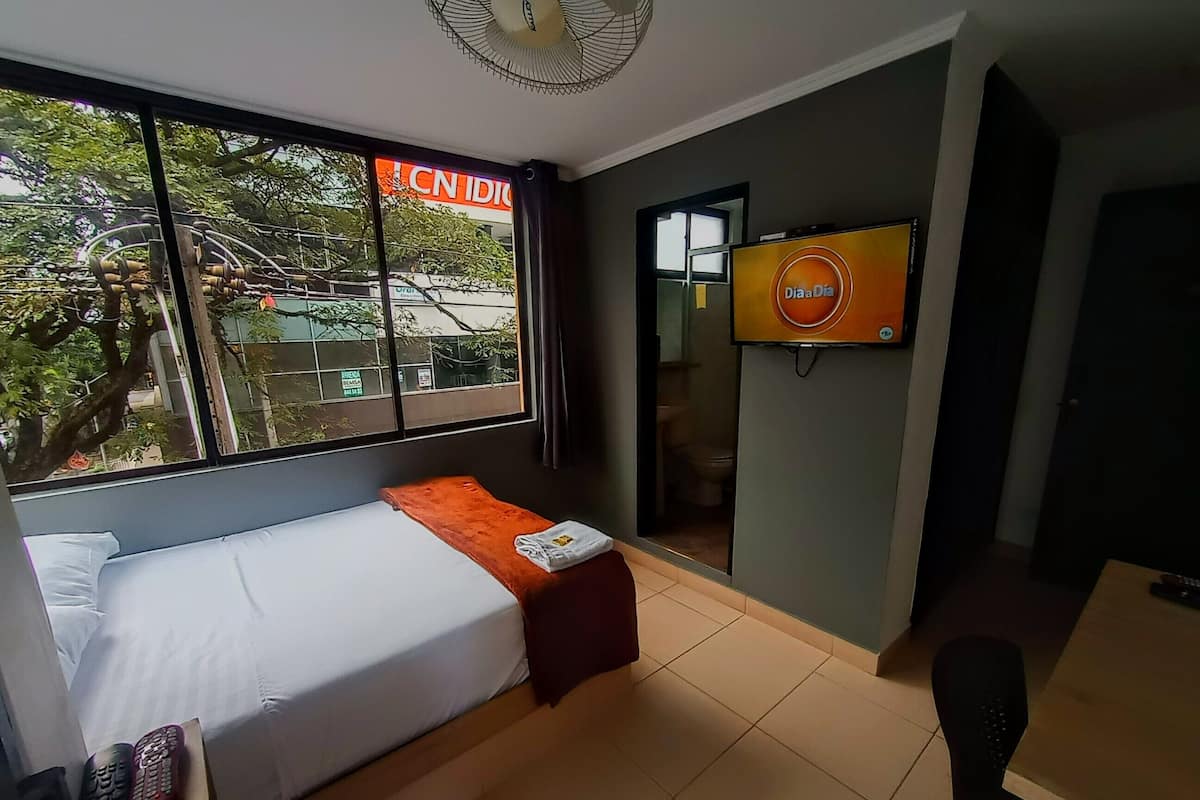Une chambre d’hôtel avec un lit, une télévision, une fenêtre donnant sur des immeubles et une petite table.
