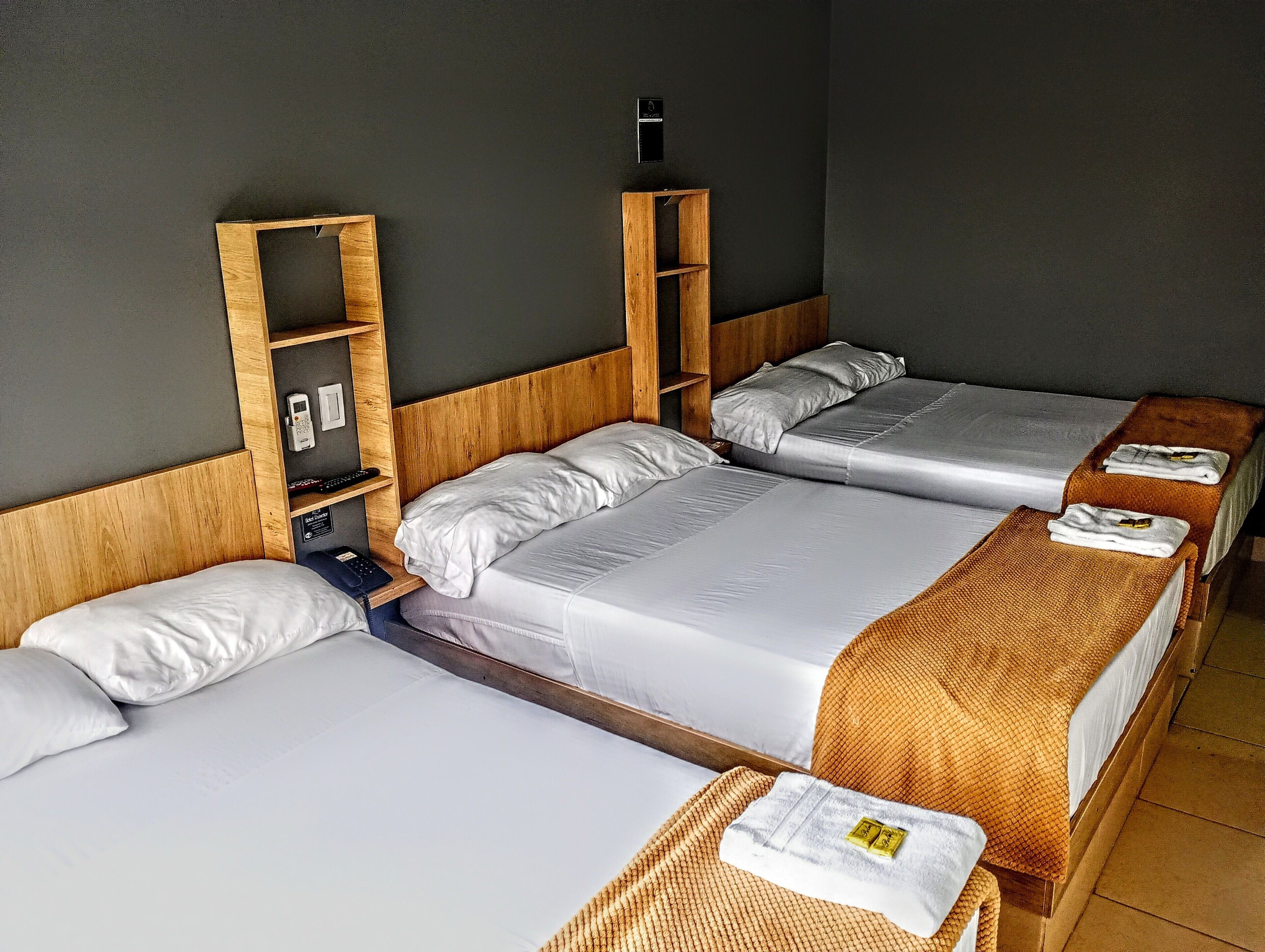 A hotel room with two single beds, a wooden headboard, a bedside table with a phone and remote control, and a small shelf with a clock.