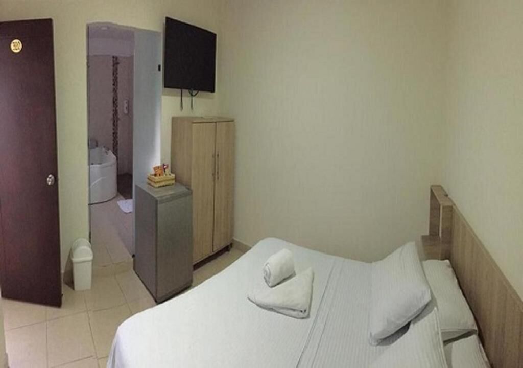 Quarto de hotel com uma cama, televisão fixada na parede, guarda-roupa e uma pequena escrivaninha com um cesto de lixo.
