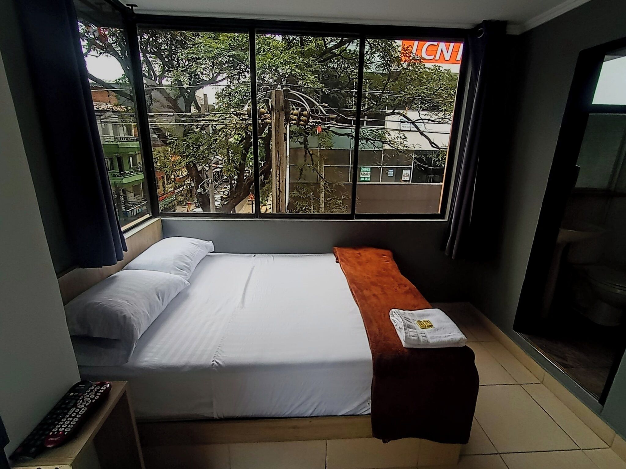 Une petite chambre d’hôtel avec un lit, un banc, une télécommande et une fenêtre donnant sur des immeubles et des arbres.