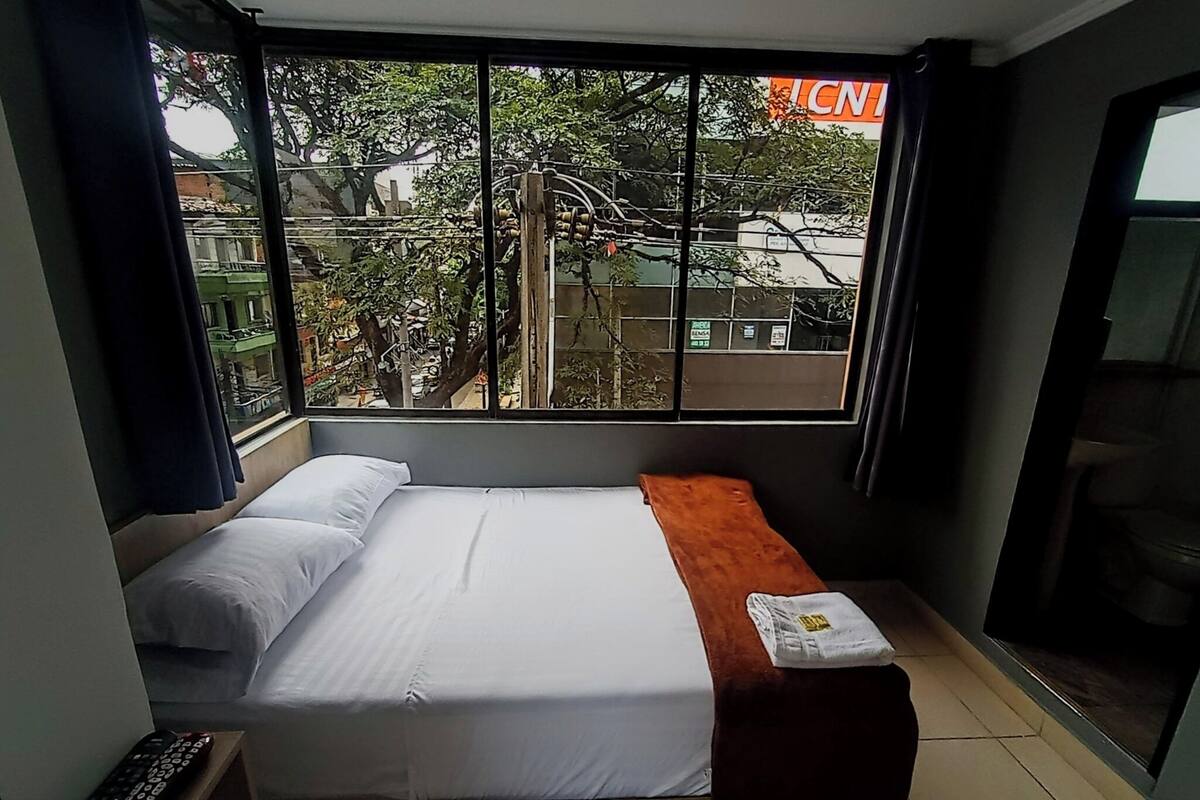 Une petite chambre d’hôtel avec un lit, un banc, une télécommande et une fenêtre donnant sur des immeubles et des arbres.