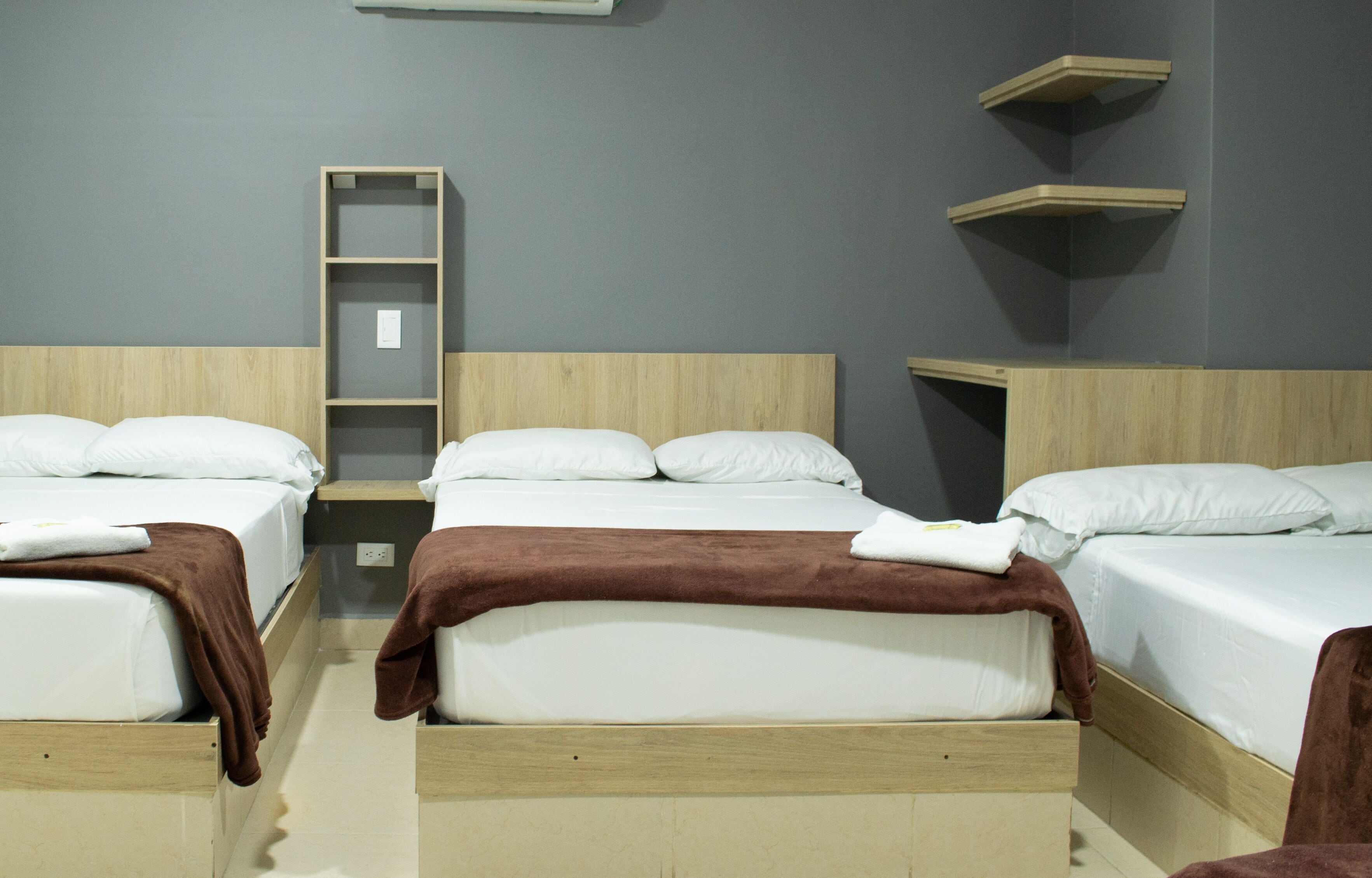 Habitación de hotel con tres camas individuales, cada una con cabecero de madera y ropa de cama blanca. Se observa una estantería de madera en la pared y una pequeña escalera de madera.