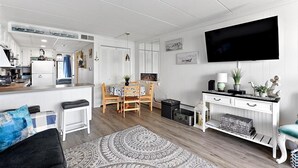 A modern kitchen with white cabinetry, a dining area with a wooden table and chairs, a living room with a sofa and a TV, and a small side table with decorative items. - Seven Seas - Recently Remodeled Ocean Block 1 Bedroom! (Ocean City)