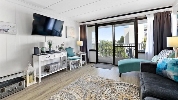 A modern living room with a flat-screen TV, a white console table, teal armchairs, a patterned rug, and a balcony with a view. - Seven Seas - Recently Remodeled Ocean Block 1 Bedroom! (Ocean City)