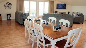 Amplia sala de estar con una mesa de comedor de madera, sillas blancas y un sofá gris.