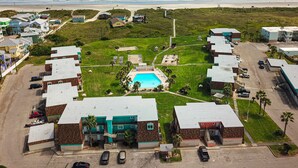 An aerial view of a coastal residential area with a swimming pool, multiple buildings, and a beach in the background. - Walk to Sandfest,  Shared Pools, Boardwalk to the Beach (Port Aransas)