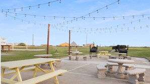 An outdoor patio area with picnic tables and stone tables, string lights overhead, and a barbecue grill in the background. - Walk to Sandfest,  Shared Pools, Boardwalk to the Beach (Port Aransas)