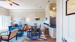 Ein Wohnzimmer mit blauem Sofa, einem Esstisch mit Stühlen, einer Küchenzeile mit Arbeitsplatte und Barhocker sowie einem Deckenventilator.