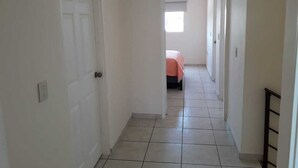A narrow hallway with white walls and tiled flooring leading to a bedroom with a bed and a window. - Casa Vacacional en la Playa (Mazatlán)
