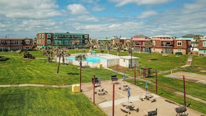 A pool area with lounge chairs and umbrellas, surrounded by green lawns and buildings with multiple stories. - Walk to Sandfest, Shared Pools, Boardwalk to Beach (Port Aransas)